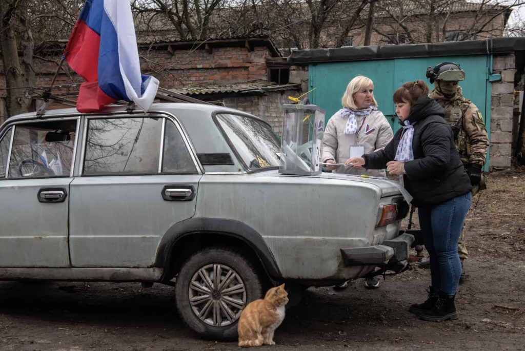 Ուկրաինայի բռնակցված տարածքների բնակիչները մարտի 10-ից մասնակցում են ՌԴ նախագահական ընտրությունների նախնական քվեարկությանը։ Մարտի 14-ին Դոնեցկում արված լուսանկարում երևում է, թե ինչպես է տեղացի կինը քվեարկում իր տան հարևանությամբ՝ Ռուսաստանի կողմից նշանակված ընտրական հանձնաժողովի անդամի և ռուս զինվորի ուղեկցությամբ: AFP/Scanpix/LETA