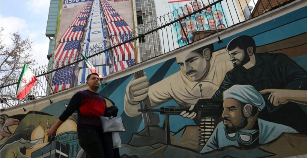 Житель Тегерана проходит мимо граффити с иранскими военными и антиамериканского плаката, Иран, 29 января 2026 года. Фото: Abedin Taherkenareh / EPA